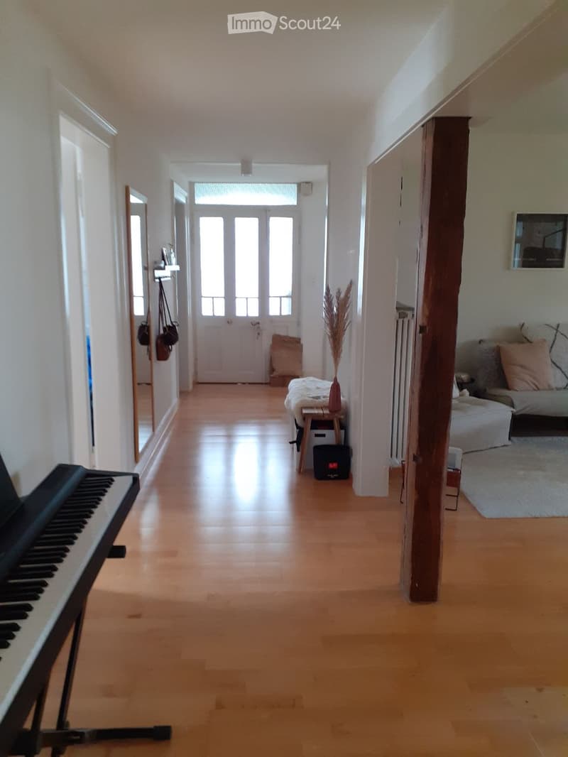 Bright hallway with wooden flooring, leading to a door at the end. Visible items include a piano on the left, a small table with a vase, and a sofa visible on the right.