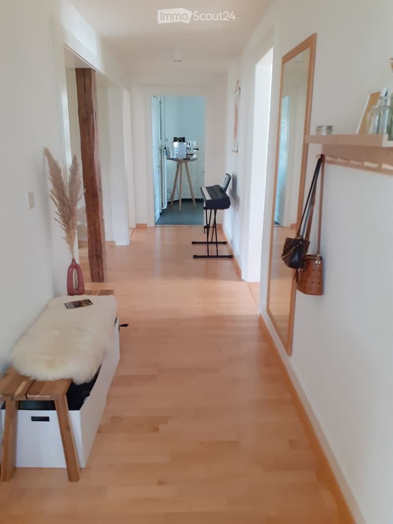 A modern hallway with light wooden flooring, white walls, and a bench with a fur throw. A small stool with glasses and a piano in the background, along with a mirror and a handbag hanging on the wall.