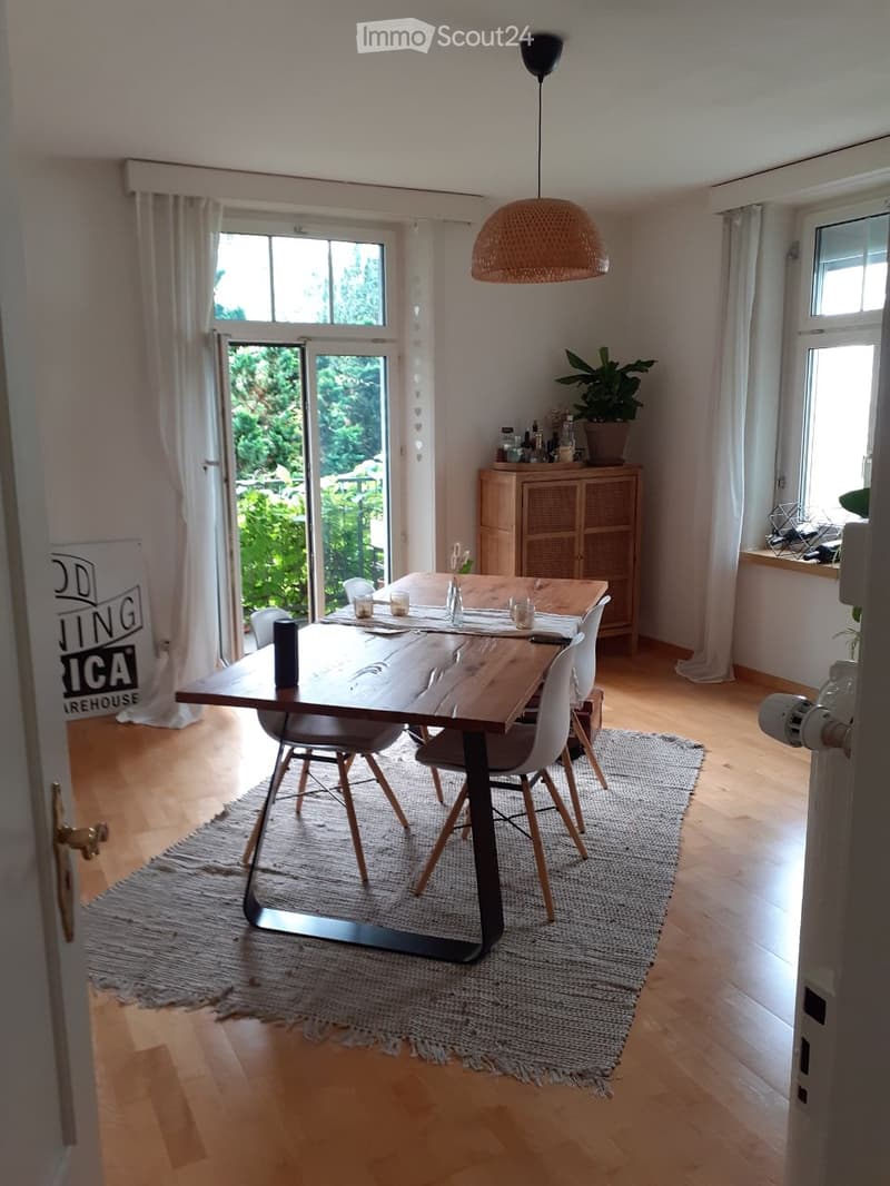 Bright dining area with wooden floor, large window, and sliding glass door leading to outdoor space. Features a wooden dining table with chairs, a woven area rug, and a hanging pendant light.