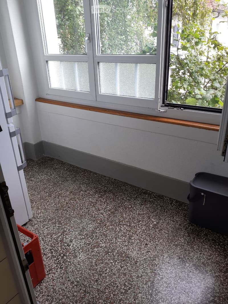Empty room with speckled gray and white speckled floor, white walls, large windows with wooden sills, and a small black storage box in the corner.