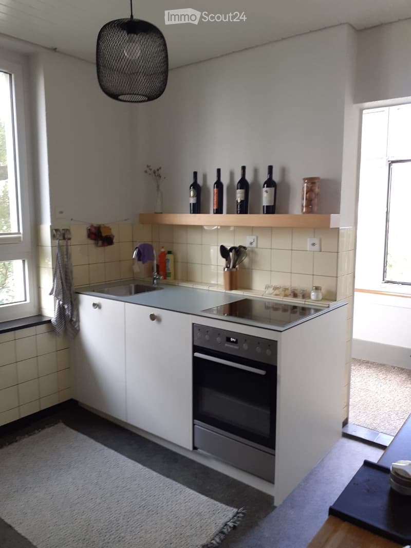 Modern kitchen with white cabinets, stainless steel sink, built-in oven, and wooden shelf holding wine bottles and decorative items.