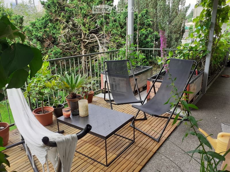 Balcony with wooden decking, two lounge chairs, a small table with drinks, and several potted plants.