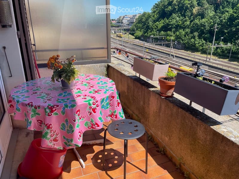 Balcony with a table covered in a pink floral tablecloth, a stool, and several potted plants, overlooking a railway track and greenery.
