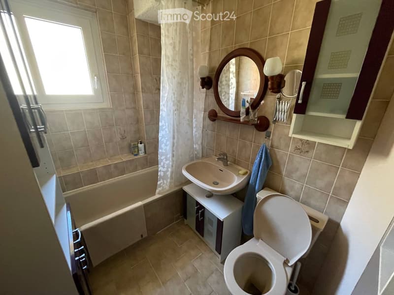 Bathroom with beige tiles, bathtub, sink, toilet, mirror, and a small cabinet. A towel hangs on the side.