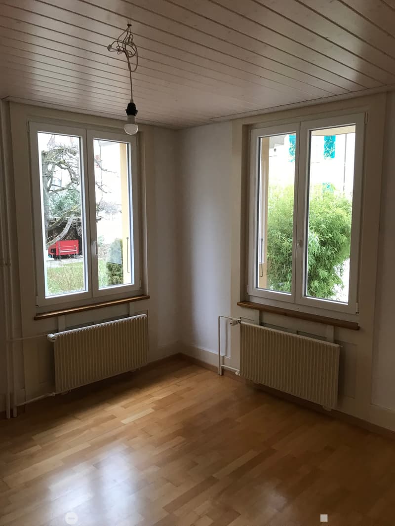 Empty room with wooden flooring, white walls, large windows, radiators, and a hanging light fixture.