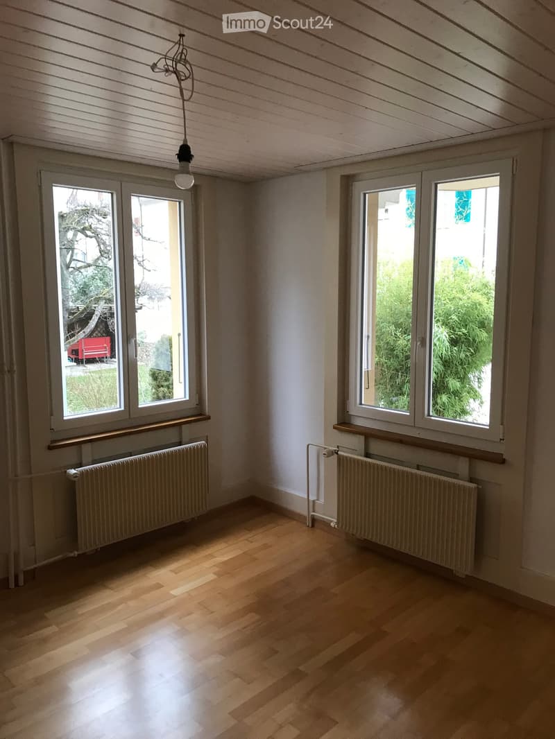 Empty room with wooden flooring, white walls, large windows, radiators, and a hanging light fixture.