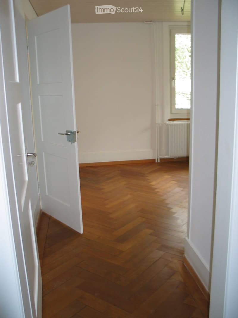 Empty room with wooden flooring and white walls, featuring a partially open door and a window with blinds.