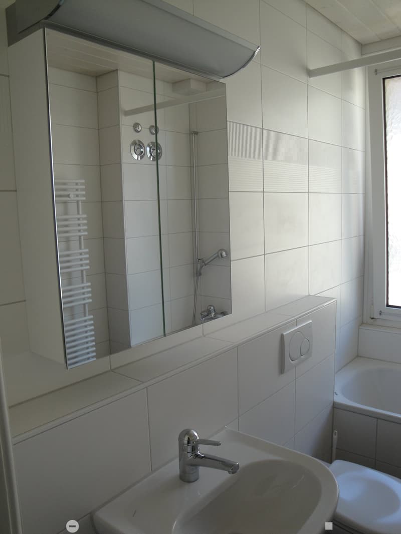 Modern bathroom with white tiles, a sink with a chrome faucet, a large mirror with built-in shelves, a bathtub, and a radiator.