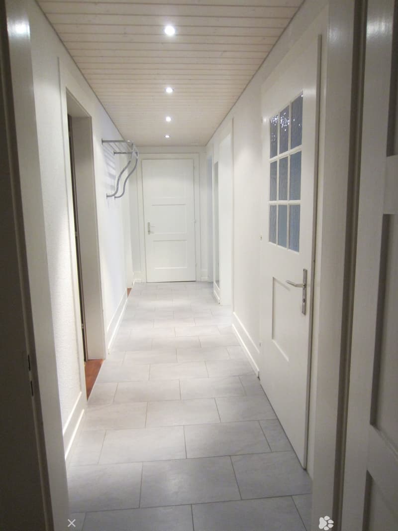 Clean, white-walled hallway with tiled flooring, recessed ceiling lights, and several doors leading to other rooms.