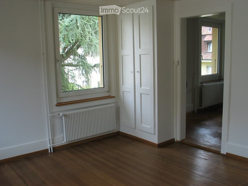 Empty room with wooden flooring, white walls, a window with a radiator, and an open door leading to another room.