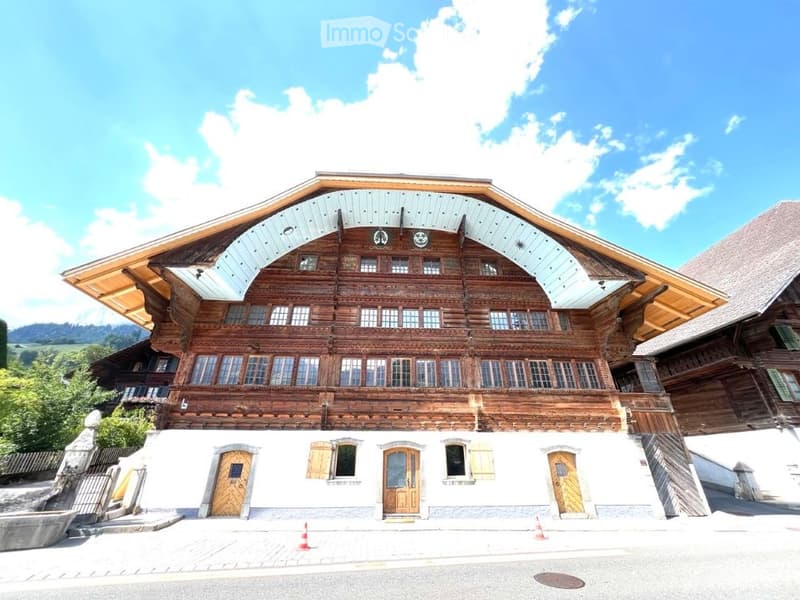 Traditional wooden chalet with a steep, multi-pitched roof and large windows, featuring a raised ground floor and multiple entrances.