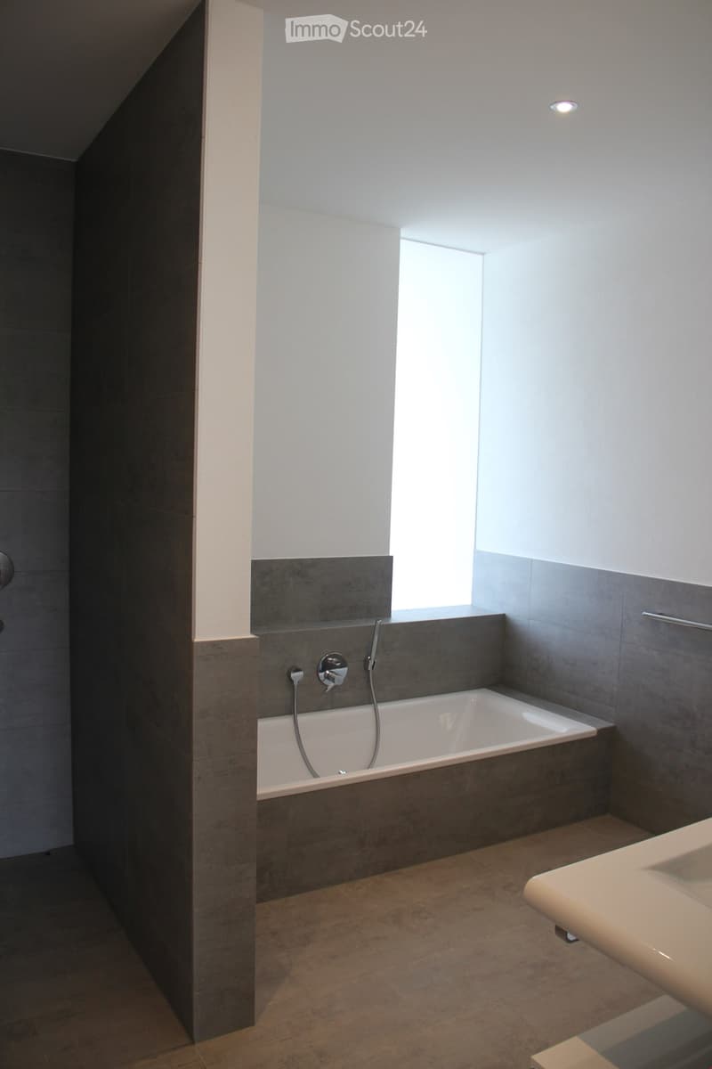 Modern bathroom with a bathtub, faucet, and sink. The walls are tiled in a combination of light and dark gray, and there is a window providing natural light.