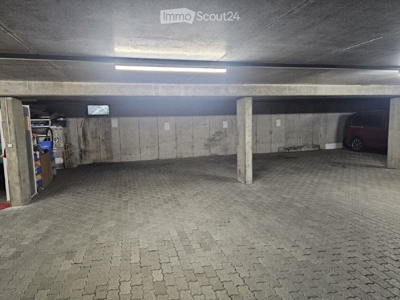 Underground parking area with concrete pillars, tiled floor, and storage shelves on the left side. A parked van is visible on the right.