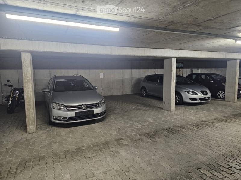Underground parking area with several parked cars and a motorcycle, concrete pillars, and a tiled floor.