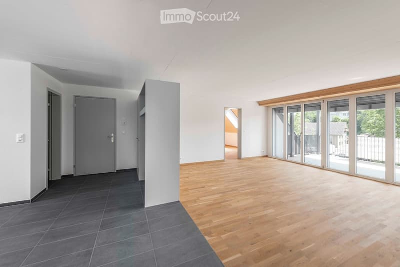 Empty room with wooden flooring, grey tiled section, large sliding glass doors leading to a balcony, and neutral white walls.