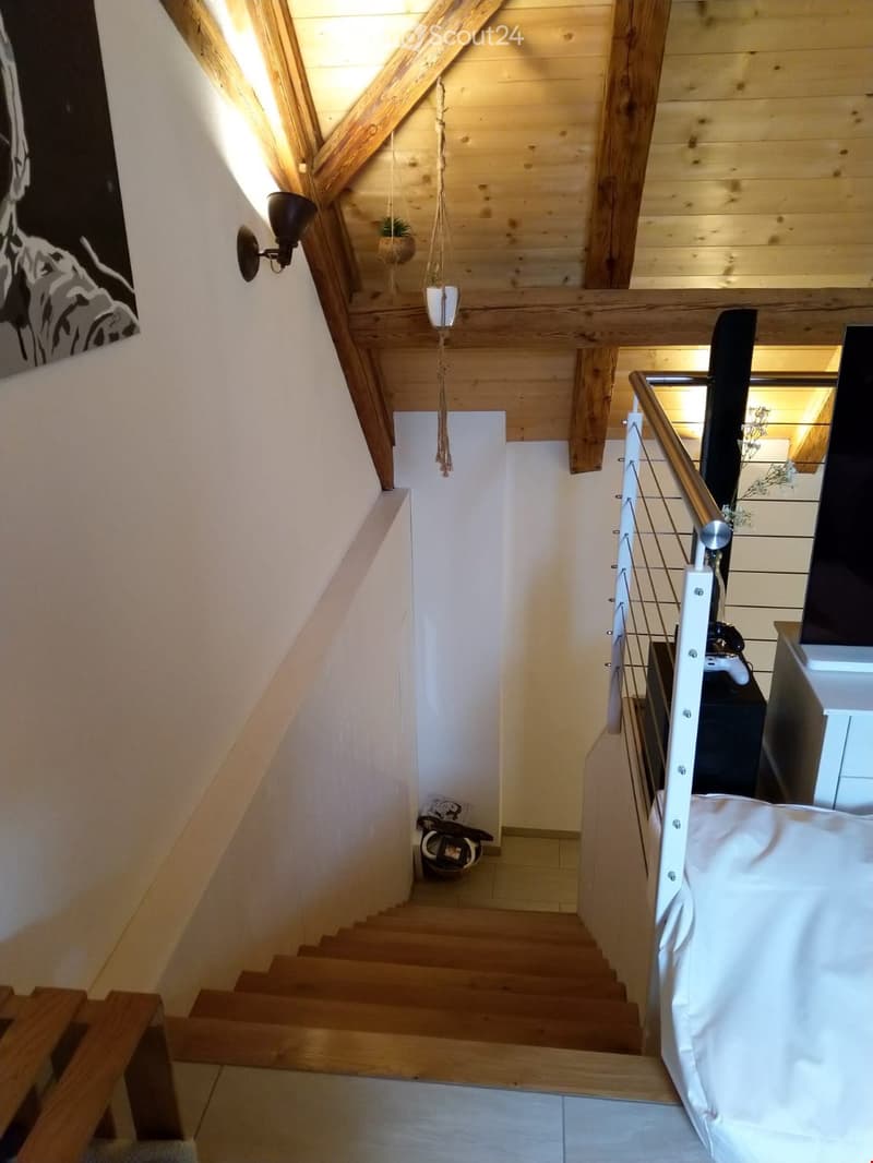 Wooden staircase with white railing, wooden walls, and hanging plants. A black wall-mounted light fixture and a black artwork are visible on the left wall. A small basket is placed at the bottom of the stairs.