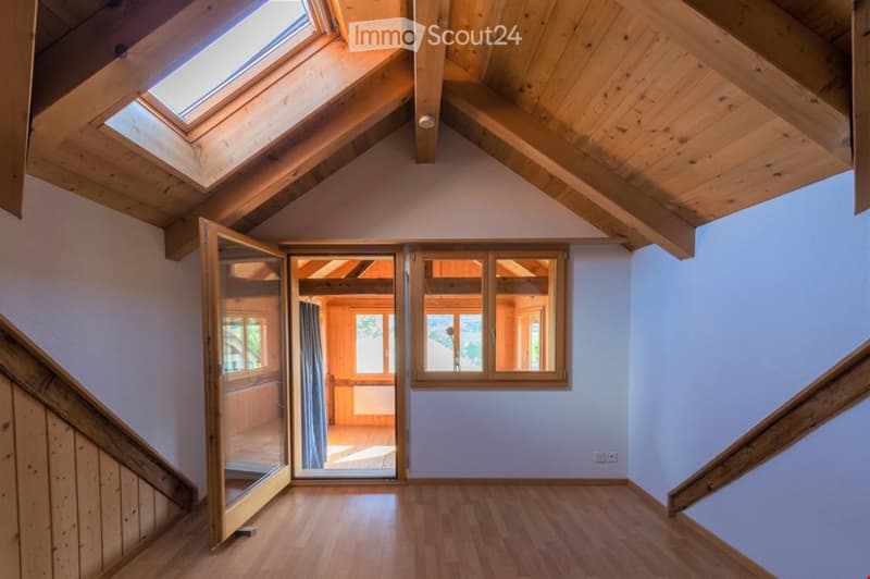 Bright, empty attic room with wooden beams, skylights, and large windows. The space features a wooden floor and white walls, with a door leading to another room.