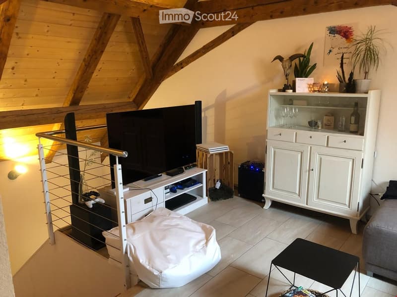 Modern living room with exposed wooden beams, a large TV on a stand, a white cabinet, a beige chair, a small black table, and a skull decoration on the shelf.