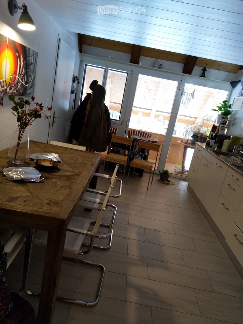 The image shows a modern, open-plan living and dining area with a wooden dining table, chairs, and a kitchen area with cabinets and a sink. There is a vase with flowers on the table, and a coat hanging on the wall. The space has large windows and a high c