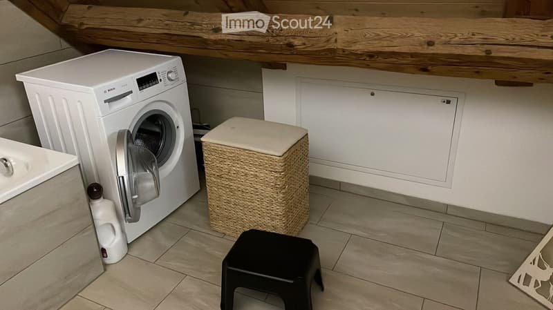 A small utility room with a white washing machine, a woven trash bin, a black plastic stool, and a bottle of cleaning supplies on the floor.