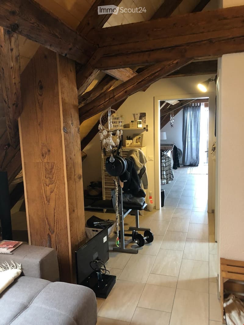 A cozy attic space with wooden beams, a grey sofa, a TV screen, an exercise bike, a dreamcatcher, and a small table with a lamp.