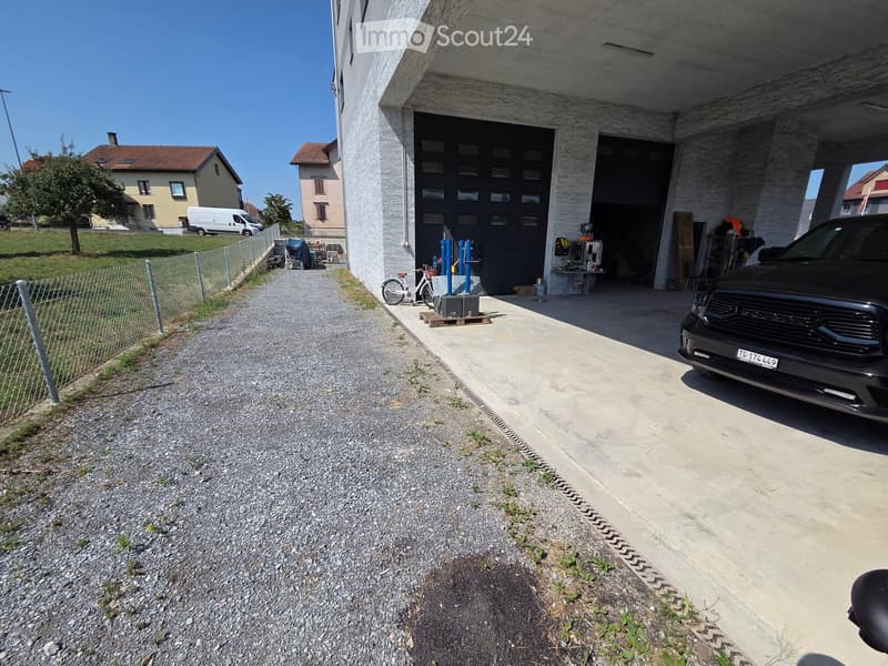 Modern garage with two large doors, a parked car, and a bicycle near the entrance. The driveway is gravel, and there is a fenced yard with a tree and neighboring houses visible in the background.