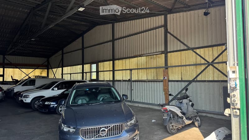 Garage with multiple parked vehicles including cars and a scooter, featuring a metal and wooden structure with large windows and a high ceiling.