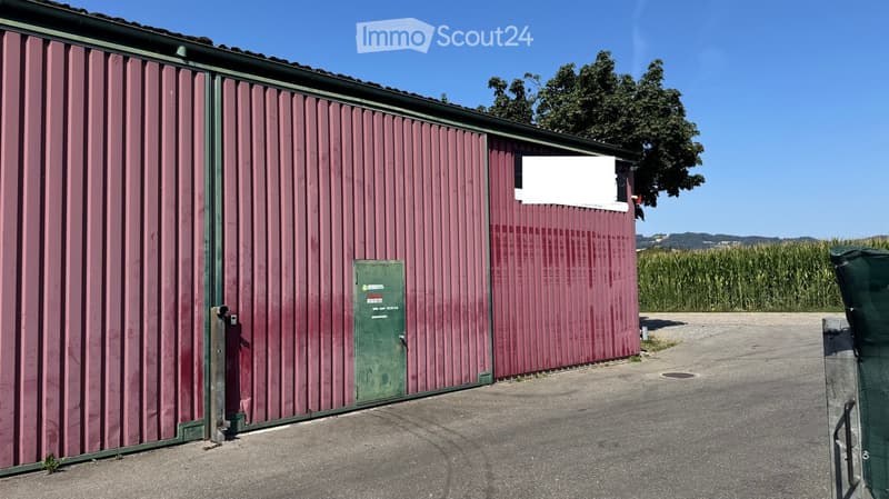 A large, industrial-style storage building with red corrugated metal walls and a green door, situated on a paved area with a cornfield in the background.