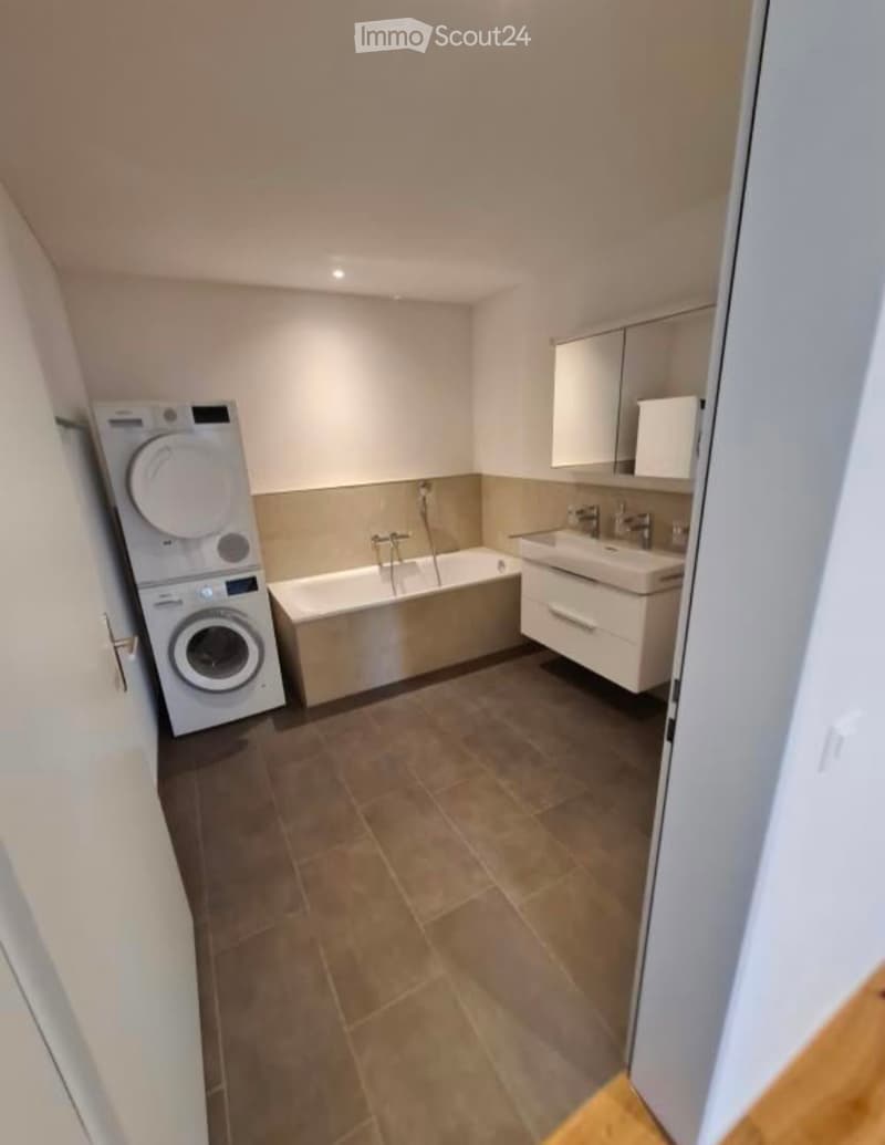 Modern bathroom with a bathtub, sink, mirror, and a private washing machine. The space features tiled flooring and neutral-colored walls.