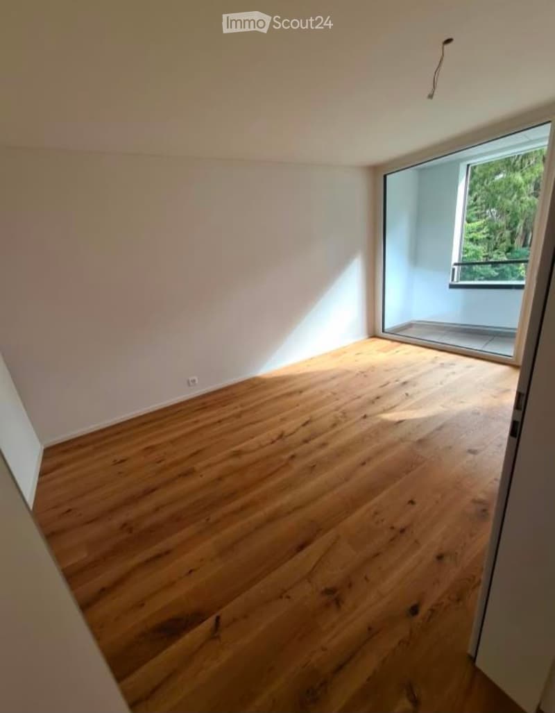 Empty room with wooden flooring and white walls, featuring a large window with a view of greenery.