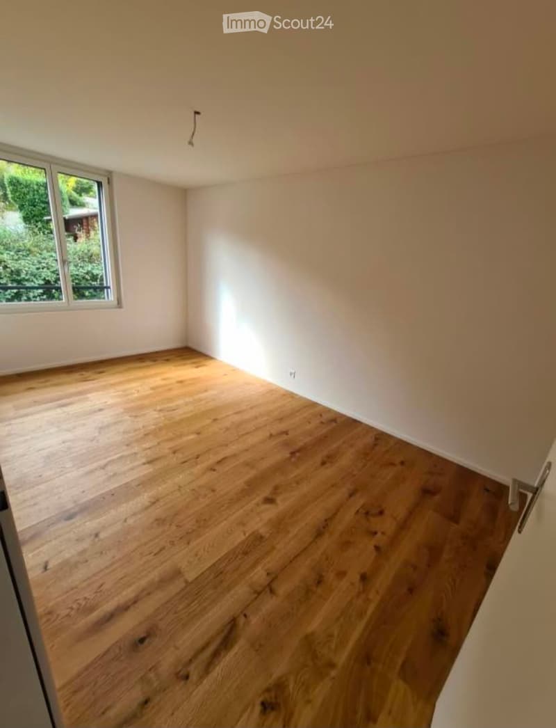 Empty room with wooden flooring and white walls, featuring a single window with a view of greenery outside.