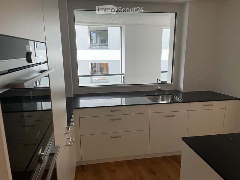 Modern kitchen with light wood cabinets, black countertops, stainless steel sink and faucet, and a large window offering a view of the building outside.