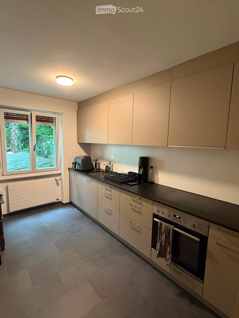 Modern kitchen with light-colored cabinets, stainless steel appliances, and a black countertop. Features an oven, coffee maker, and a sink with a dish rack.