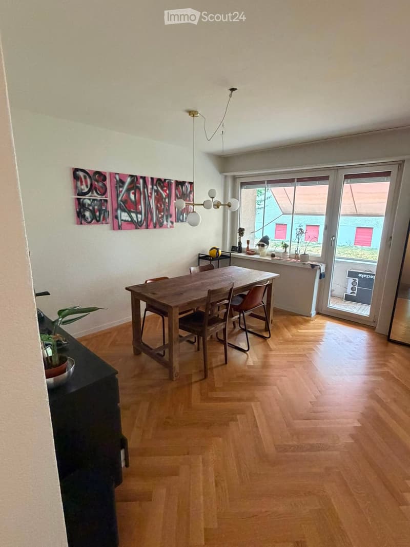 Dining room with wooden floor, dining table, chairs, potted plant, artwork, and a view through a large window.