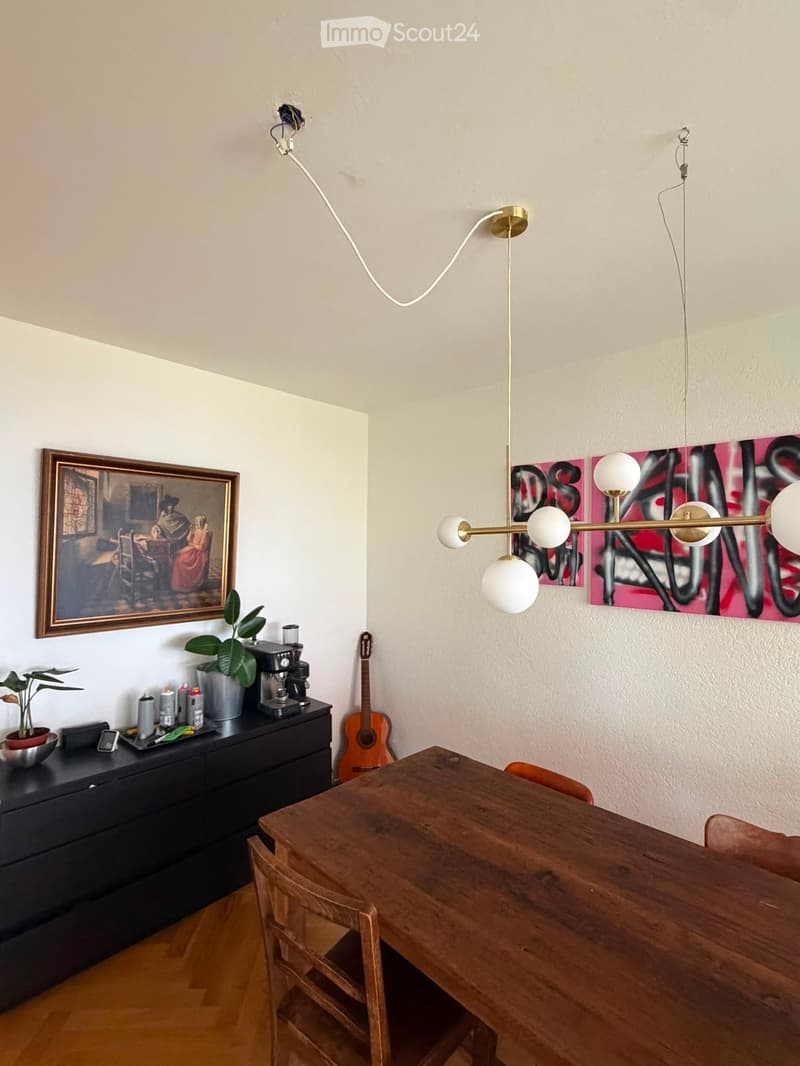 A modern, well-lit room with a wooden dining table and chairs, a black cabinet with a coffee maker, a guitar, and a decorative lamp hanging from the ceiling.