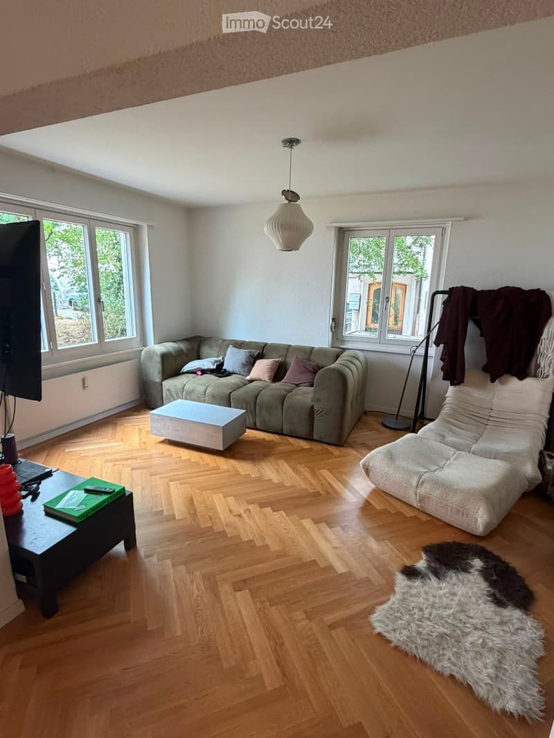 Living room with wooden flooring, large windows, a green sofa, a white coffee table, a white chair, a black table with a green book, a floor lamp, and a fur rug.