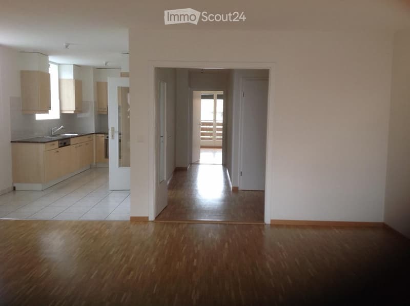 Empty hallway with wooden flooring, leading to a kitchen area with light wood cabinets, a sink, and a dishwasher. The hallway has white walls and a door leading to another room.