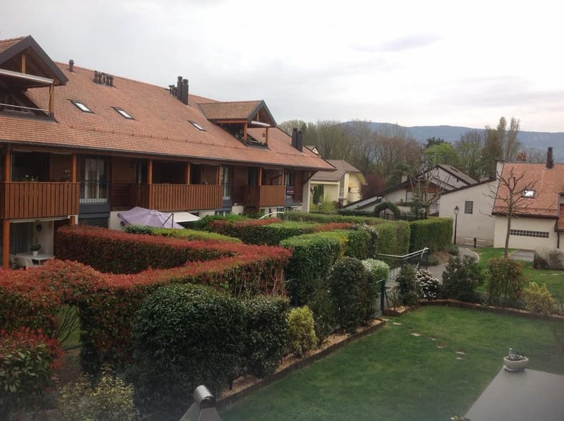 Well-maintained garden with neatly trimmed hedges, a small patio area, and a wooden deck on the house. The house features a red-tiled roof and large windows, with a scenic mountain view in the background.