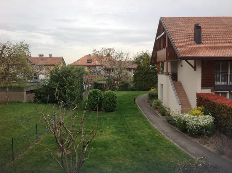 Well-maintained garden with green lawn, trimmed bushes, small tree, and pathway leading to a house with a wooden staircase and balcony.