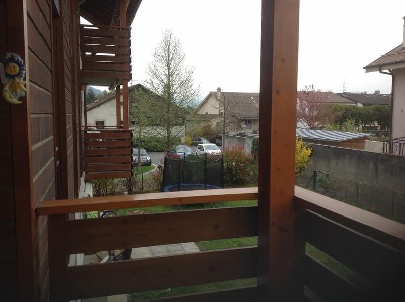 Wooden balcony overlooking a residential area with houses, trees, parked cars, and a small trampoline in the yard.