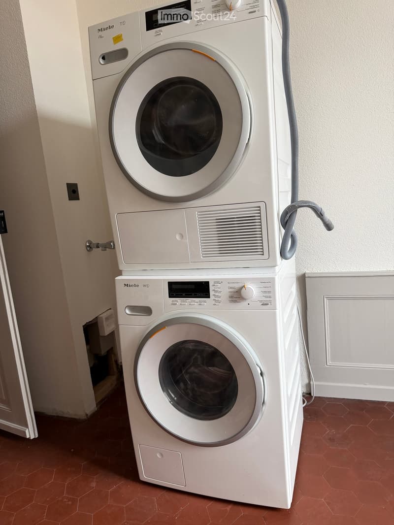 A compact bathroom with a stacked Miele washing machine and dryer unit, featuring a control panel and a hose connection.
