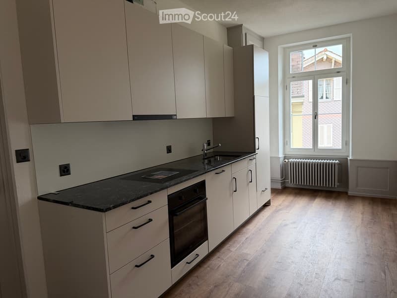 Modern kitchen with light-colored cabinets, a black countertop, a sink, and an oven. The room features wooden flooring and a large window with a view of the street.
