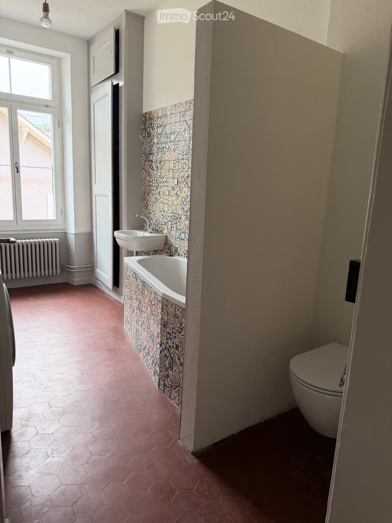 Bathroom with hexagonal pink tiles, a sink, bathtub, and toilet. The walls feature decorative tiles with geometric patterns. There is a window with a view of a roof and a radiator below it.