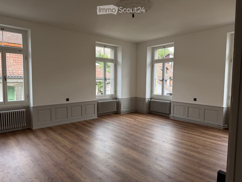 Empty room with wooden flooring, light-colored walls, multiple windows, radiators, and a decorative ceiling light fixture.