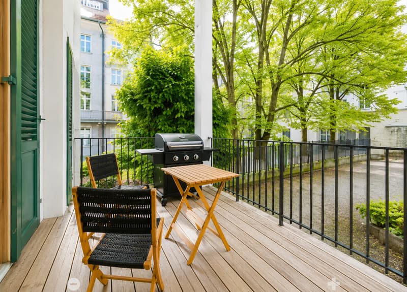 Balcony with wooden decking, black metal railing, small wooden table, two black wicker chairs, and a grill.