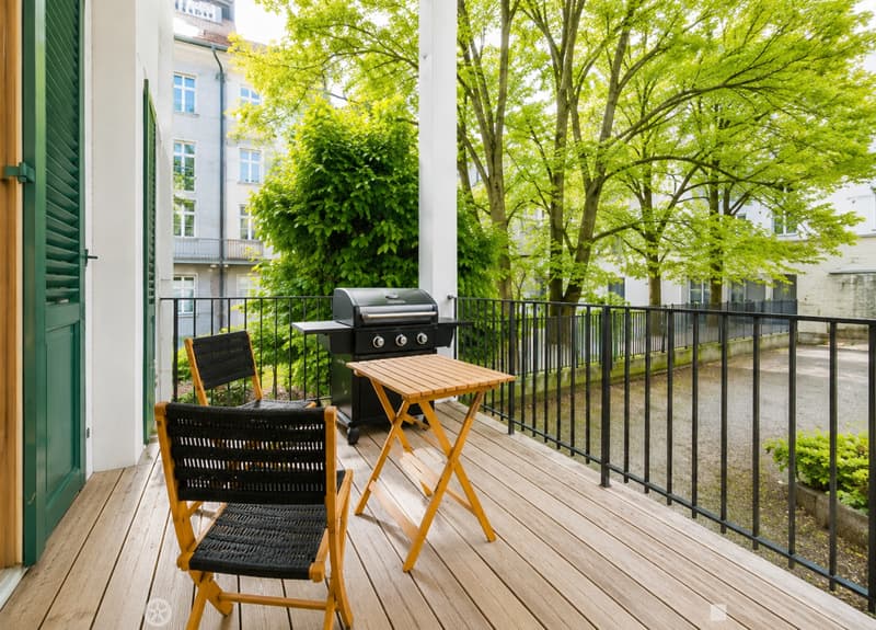 Balcony with wooden decking, black metal railing, small wooden table, two black wicker chairs, and a grill.