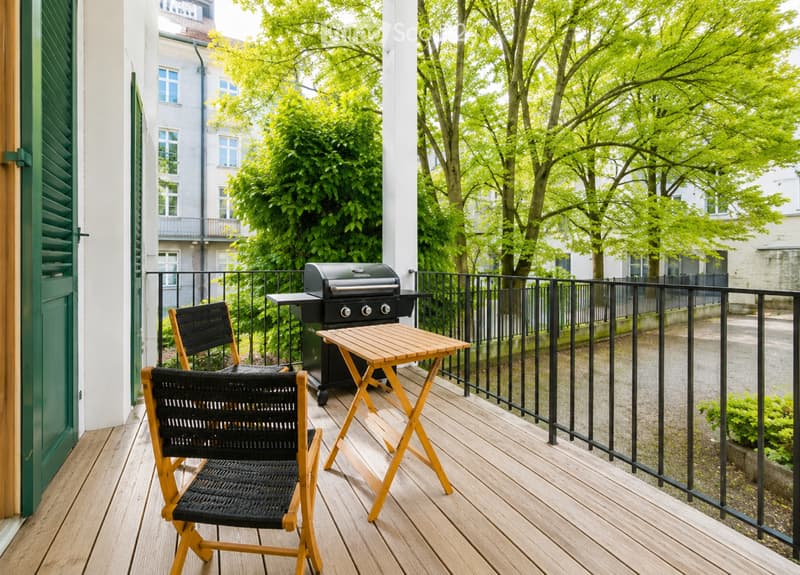 Balcony with wooden decking, black metal railing, small wooden table, two black wicker chairs, and a grill.