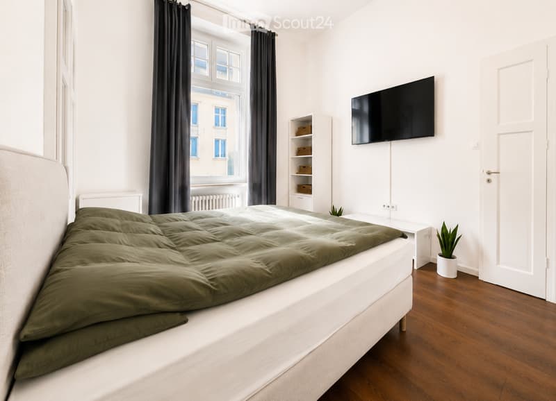 A modern bedroom with a large bed, green duvet, nightstand, television mounted on the wall, and a small potted plant on the floor.