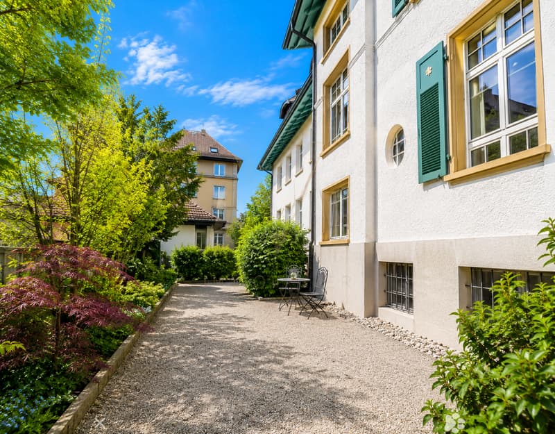 A gravel pathway leads to a small seating area with a table and two chairs, surrounded by lush greenery and a white building with green shutters.