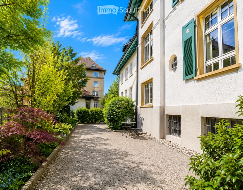 A gravel pathway leads to a small seating area with a table and two chairs, surrounded by lush greenery and a white building with green shutters.